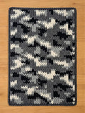 A chunky knit blanket in a winter camouflage pattern with black, grey, and cream yarn, displayed flat on a light wood floor.