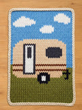 A knitted chunky blanket featuring a camper van design. The blanket has a blue sky with white clouds at the top, a beige camper van in the middle, and green grass at the bottom. The blanket is bordered with a cream-colored knitted edge.