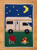 A knitted camper scene with a blue night sky, yellow stars, and a crescent moon. The camper is white with black wheels, and a red picnic table and campfire are in the foreground. The border is cream-colored.