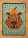 A knitted blanket featuring a brown capybara face with an orange fruit on its head. The blanket has a light green background and a darker green border. The capybara's face is detailed with a black smile and eyes.