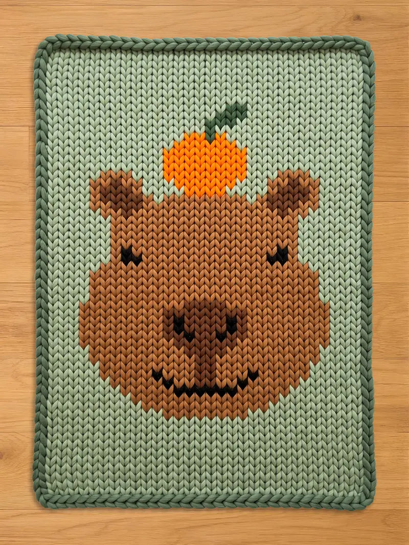 A knitted blanket featuring a brown capybara face with an orange fruit on its head. The blanket has a light green background and a darker green border. The capybara's face is detailed with a black smile and eyes.