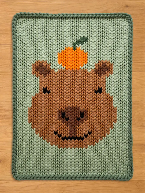 A knitted blanket featuring a brown capybara face with an orange fruit on its head. The blanket has a light green background and a darker green border. The capybara's face is detailed with a black smile and eyes.
