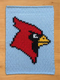 A knitted chunky blanket featuring a red cardinal bird design on a light blue background. The cardinal is rendered in a pixelated style, with black, white, and yellow accents. The blanket has a textured appearance, suggesting a handmade item.