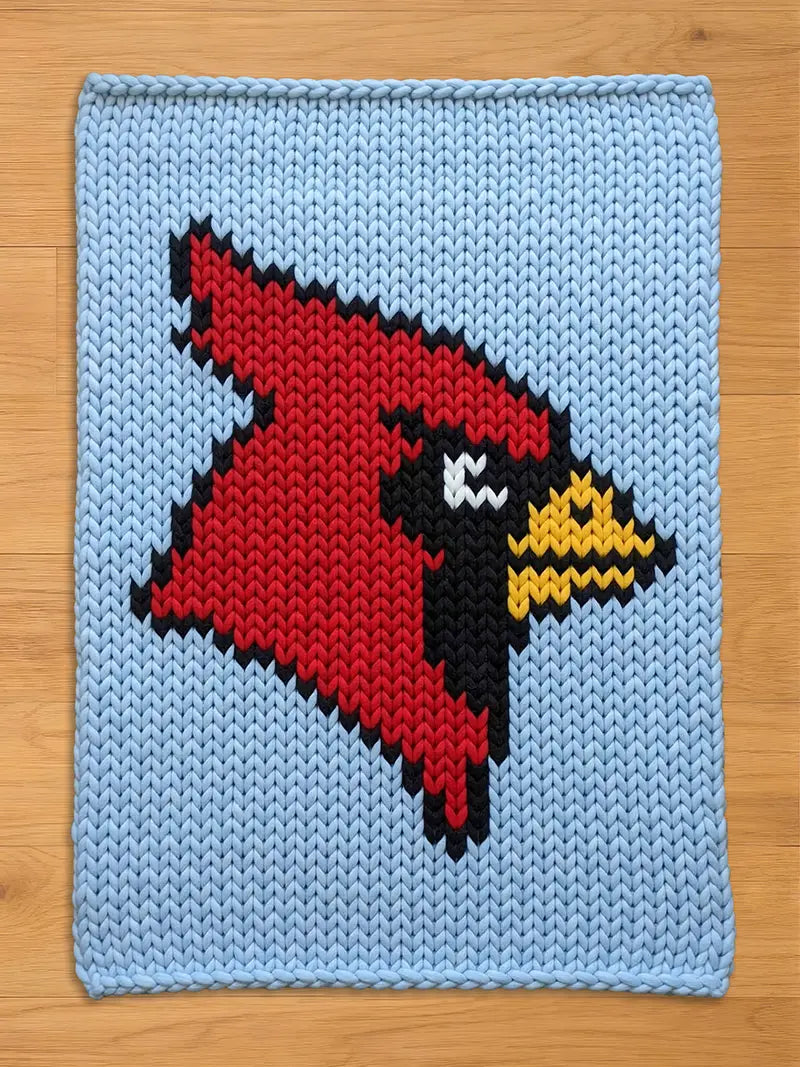 A knitted chunky blanket featuring a red cardinal bird design on a light blue background. The cardinal is rendered in a pixelated style, with black, white, and yellow accents. The blanket has a textured appearance, suggesting a handmade item.