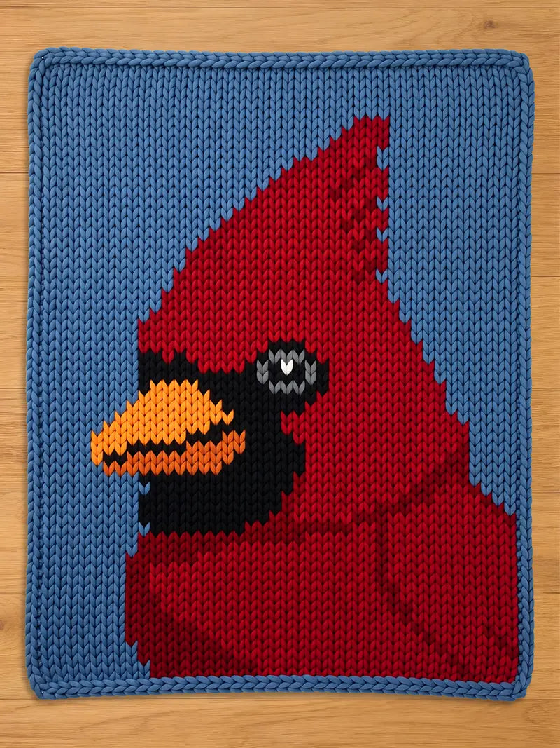 A knitted chunky knit blanket featuring a vibrant cardinal bird design. The bird is primarily red with a black face and a yellow beak, set against a light blue background. The knitted texture is clearly visible.