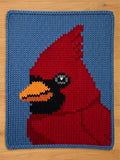 A knitted chunky knit blanket featuring a vibrant cardinal bird design. The bird is primarily red with a black face and a yellow beak, set against a light blue background. The knitted texture is clearly visible.