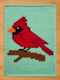 A knitted chunky knit blanket featuring a red cardinal bird perched on a brown branch against a light green background. The bird has black accents and an orange beak. The piece is made of yarn.