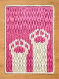 A rectangular, knitted chunky blanket in shades of pink and white. The blanket features a repeating pattern of two stylized, white objects with pink accents against a pink background. The blanket is bordered with a white knitted edge.