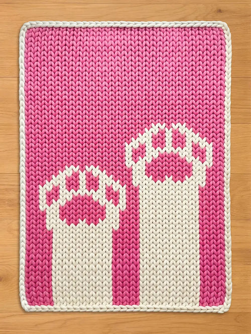 A rectangular, knitted chunky blanket in shades of pink and white. The blanket features a repeating pattern of two stylized, white objects with pink accents against a pink background. The blanket is bordered with a white knitted edge.