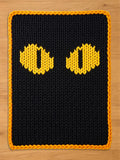 Chunky knit blanket featuring a black background with large, bright yellow cat eyes in a vertical pupil pattern, finished with an orange braided border, laid flat on a light wood floor.