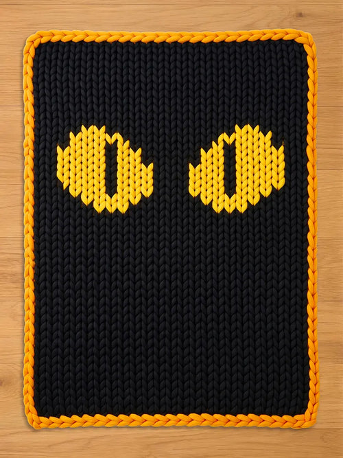 Chunky knit blanket featuring a black background with large, bright yellow cat eyes in a vertical pupil pattern, finished with an orange braided border, laid flat on a light wood floor.