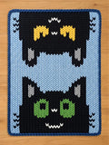 A knitted chunky blanket featuring a stacked cat design. The bottom cat has green eyes and a white mouth, while the top cat has yellow ears. The background is black with a light blue border and a dark blue trim.
