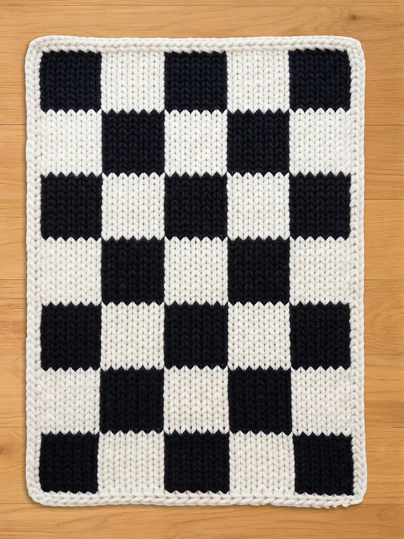 Knitted checkerboard pattern in black and white. The design features alternating black and white squares, with a white border. The item is likely a blanket or a decorative piece for the home.