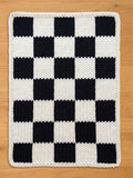 Knitted checkerboard pattern in black and white. The design features alternating black and white squares, with a white border. The item is likely a blanket or a decorative piece for the home.