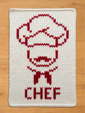 White chunky knit blanket featuring a red knit outline of a chef wearing a chef's hat and mustache, with the word "CHEF" knitted in red at the bottom, displayed on a wooden floor.