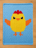Handcrafted chunky knit blanket in light blue featuring a large, pixel-art style yellow baby chick pattern with orange wings, an orange beak, and a red comb, displayed on a wooden surface.