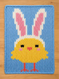 A chunky knit blanket featuring a yellow Easter chick wearing white and pink bunny ears, set against a light blue background.