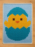 A knitted Easter-themed chunky blanket featuring a yellow chick hatching from a blue egg. The chick has brown eyes and an orange beak. The blanket has a light blue background with a white border.