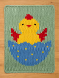  A knitted chunky blanket featuring a yellow chick hatching from a blue egg. The chick has red accents and black eyes. The egg is decorated with yellow details, all set against a green background.