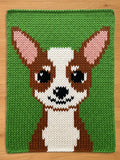 A knitted chunky blanket featuring a cartoon-style dog face. The dog has brown and white fur, black eyes, and pink ears, set against a green background. The piece is likely a decorative item or part of a larger craft project.