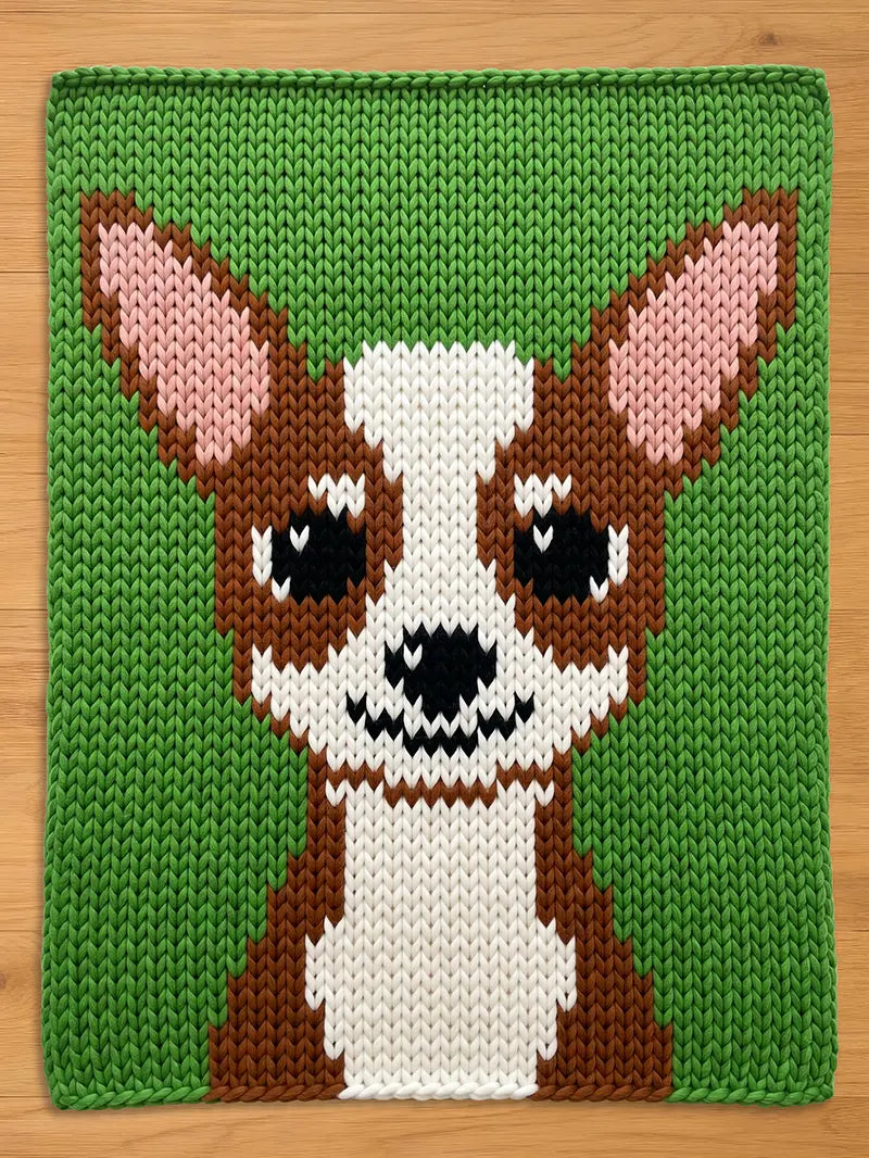 A knitted chunky blanket featuring a cartoon-style dog face. The dog has brown and white fur, black eyes, and pink ears, set against a green background. The piece is likely a decorative item or part of a larger craft project.