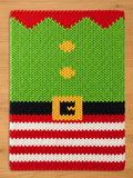 A knitted Christmas-themed rectangle chunky blanket with a green body, red collar, and red and white striped bottom. The design resembles an elf's outfit, complete with a black belt and gold buckle. Two yellow buttons are visible on the green section.