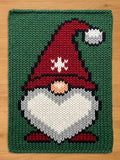 A knitted Christmas gnome design on a green background. The gnome has a red hat with white snowflake accents, a white beard, and a peach-colored nose. The design is rectangular and appears to be a rug or decorative piece.
