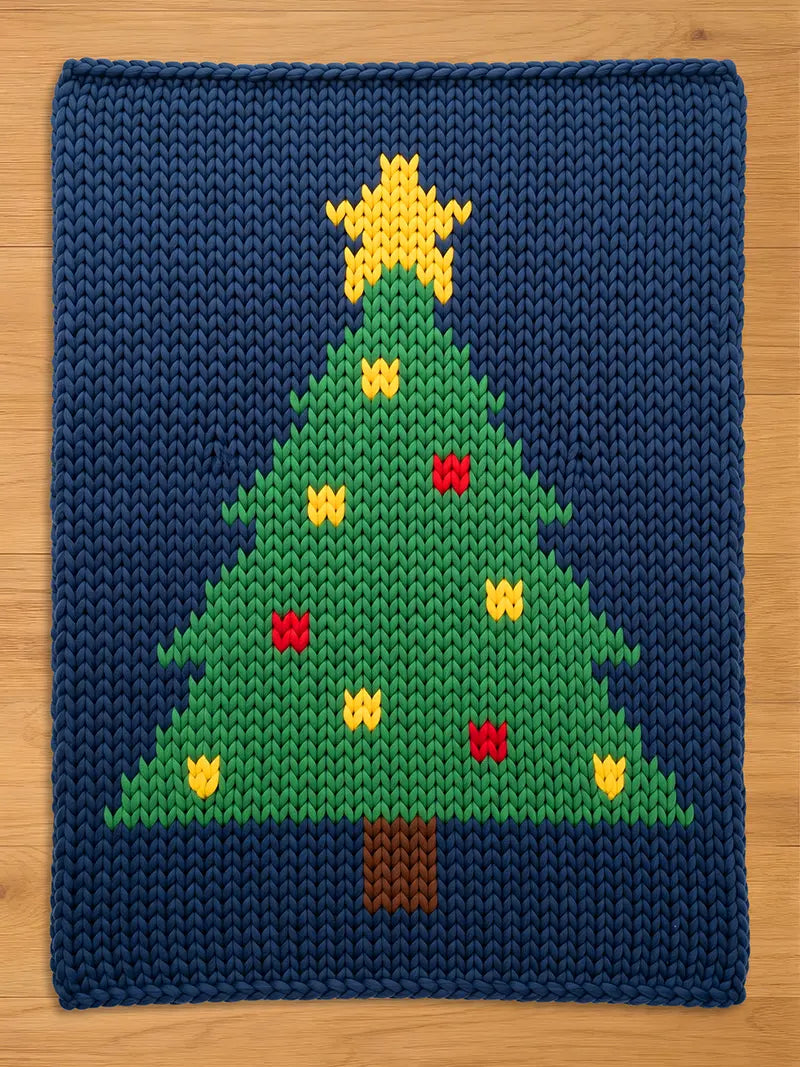 A knitted Christmas tree design on a navy blue background. The tree is green with yellow and red ornaments, a brown trunk, and a yellow star on top. The item is likely a decorative mat or blanket.