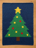 A knitted Christmas tree design on a navy blue background. The tree is green with yellow and red ornaments, a brown trunk, and a yellow star on top. The item is likely a decorative mat or blanket.