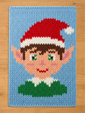 A rectangular chunky knit blanket featuring a graphic of an elf face. The elf has brown hair, green eyes, pink cheeks, and a red and white hat. The elf wears a green collar with a white trim, all set against a light blue background.