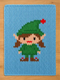 A rectangular light blue knit blanket featuring a central graphic of an elf. The elf has brown hair, a green hat with a red star, a green tunic, and brown shoes with red accents. The knit texture is consistent across the entire surface.