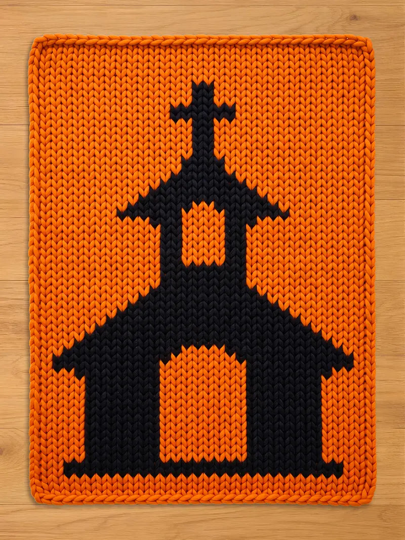 Chunky knit blanket in bright orange featuring a bold black knitted church pattern with a steeple and cross, laid flat on a light wood surface.