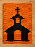 Chunky knit blanket in bright orange featuring a bold black knitted church pattern with a steeple and cross, laid flat on a light wood surface.