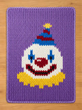 A chunky knit blanket featuring a purple background with a woven clown face pattern. The clown design includes a white face, blue eye makeup, a red nose and smile, a yellow ruffled collar, and a blue and red striped party hat.