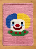 Chunky knit blanket in pastel pink featuring a large, hand-knitted clown face design in the center. The clown has split blue and yellow hair, a white face, a red nose and smile, and a green ruffled collar with red details, laid flat on a light wood surface.