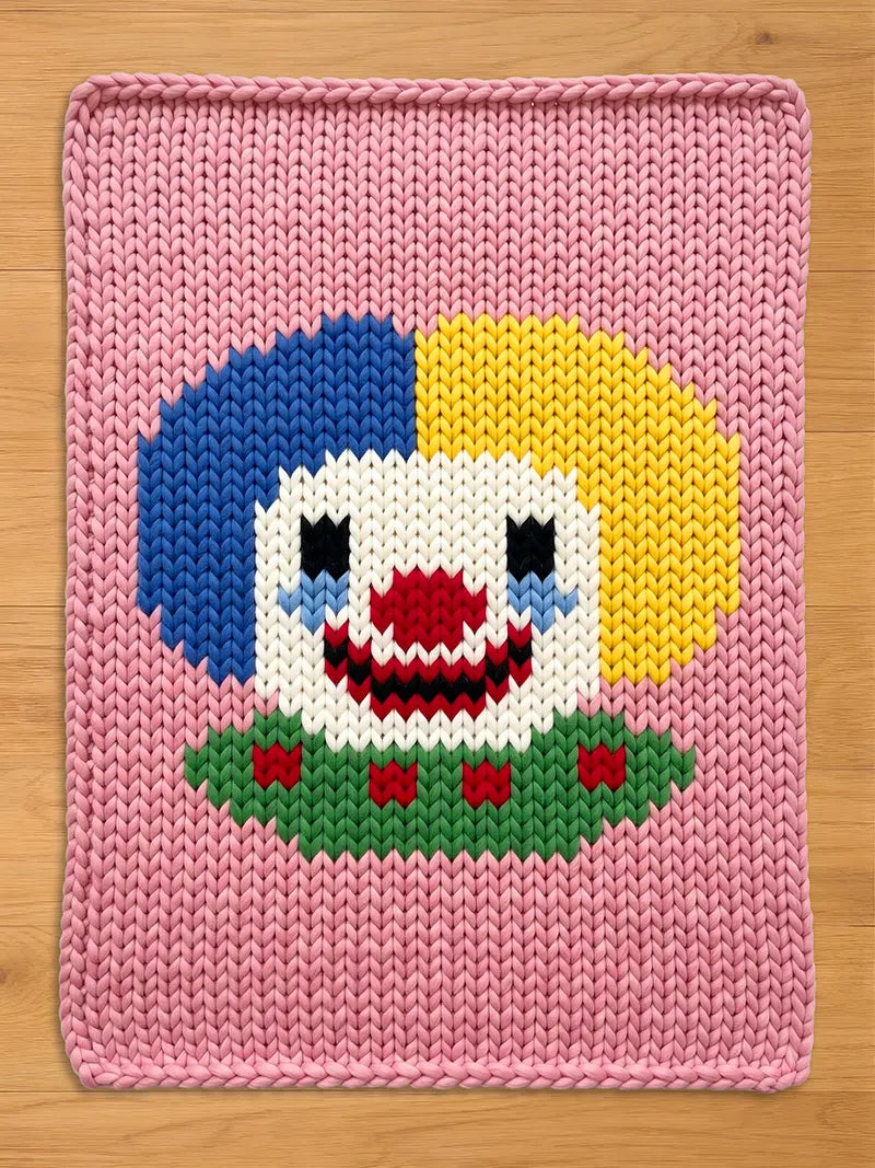 Chunky knit blanket in pastel pink featuring a large, hand-knitted clown face design in the center. The clown has split blue and yellow hair, a white face, a red nose and smile, and a green ruffled collar with red details, laid flat on a light wood surface.