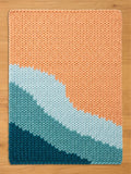 A rectangular chunky knit blanket featuring a color-blocked design with diagonal stripes. The pattern transitions from a large section of peach, through a band of light blue, to a section of teal, and finally a dark blue corner.
