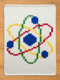 White knitted square with a colorful atom design. The atom features blue, red, yellow, and green elements, creating a vibrant and educational theme. The background is a light wood surface.
