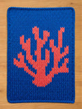 A knitted chunky blanket in a vibrant blue color. The design features a coral-like pattern in a contrasting coral-red hue. The edges are neatly finished, creating a clean, modern look.