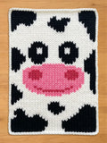 A chunky knitted cow face design with a white background and black spots. The cow's nose is pink, and the eyes are black. The edges are finished with a white border. This item is suitable for home decor.