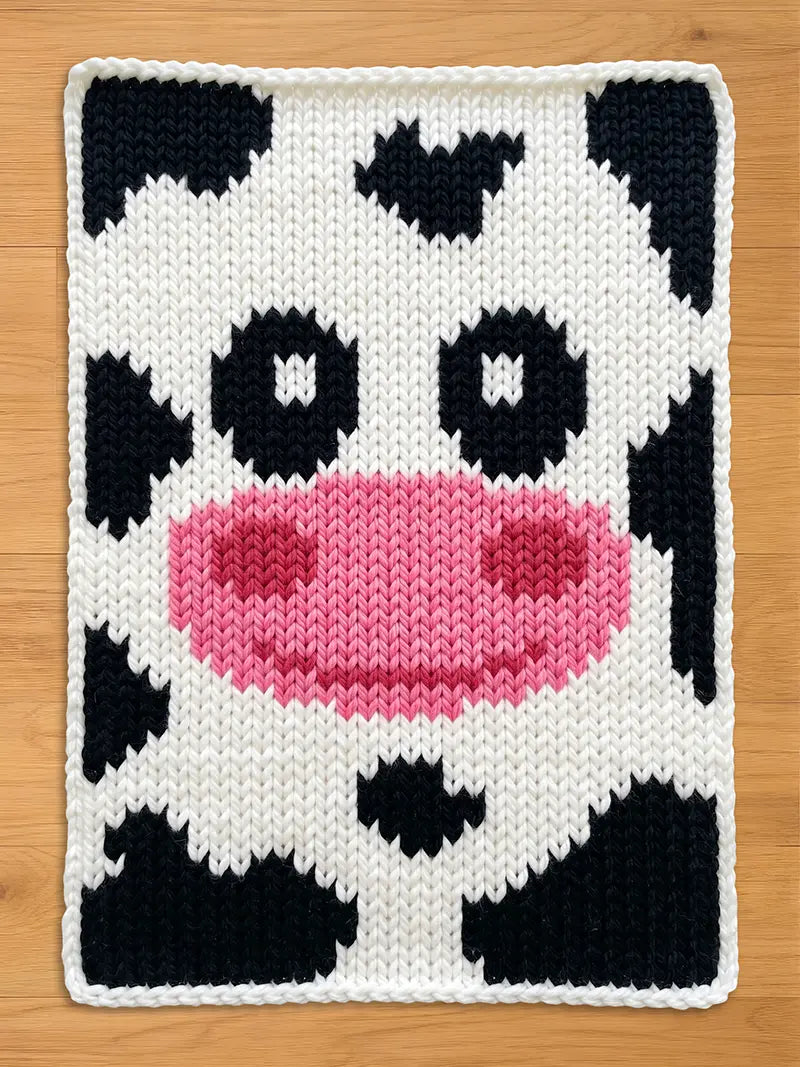 A chunky knitted cow face design with a white background and black spots. The cow's nose is pink, and the eyes are black. The edges are finished with a white border. This item is suitable for home decor.