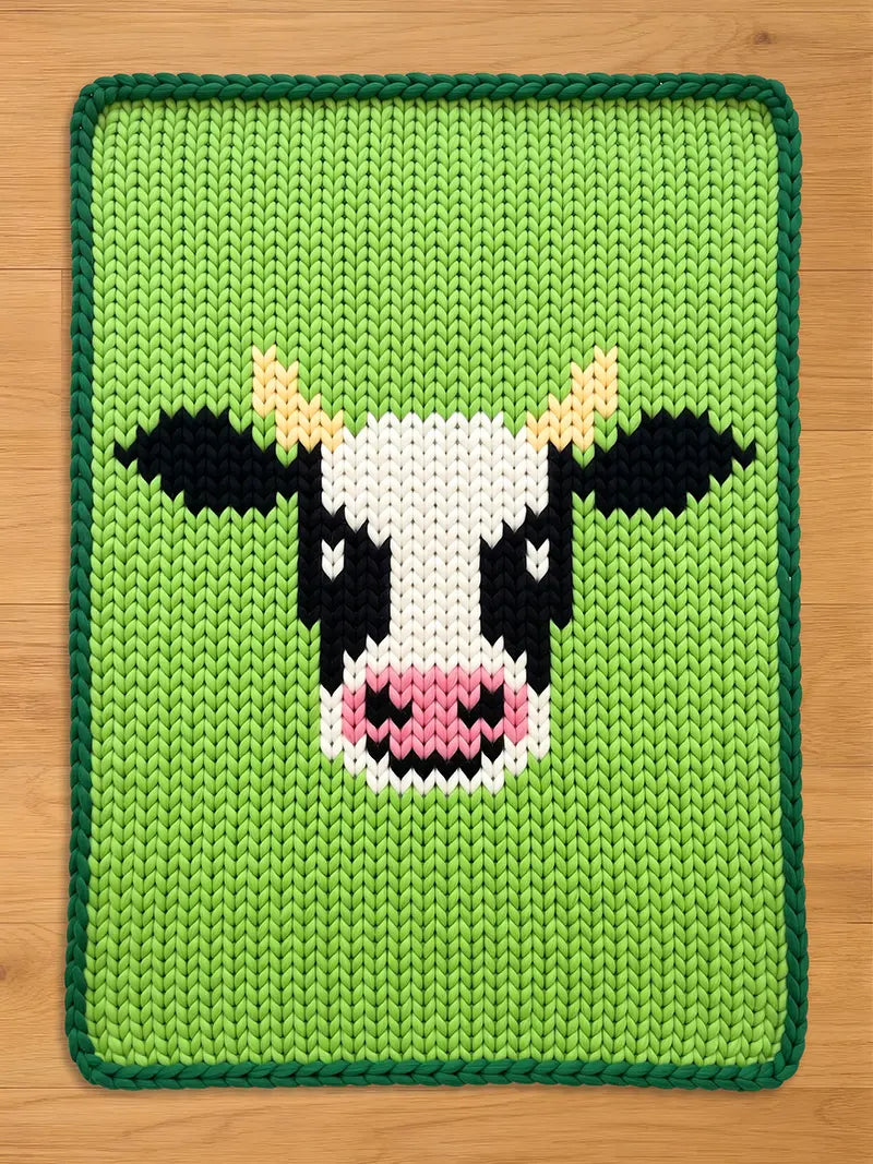 Chunky knit blanket featuring a black and white cow face pattern on a bright green background with a dark green braided border.