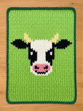 Chunky knit blanket featuring a black and white cow face pattern on a bright green background with a dark green braided border.