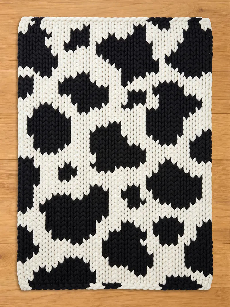 A rectangular, hand-knitted chunky blanket with a cowhide pattern. The blanket features large, irregular black shapes against a white background. The thick yarn creates a textured surface, suitable for a living room or bedroom.