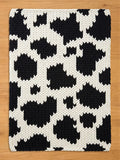 A rectangular, hand-knitted chunky blanket with a cowhide pattern. The blanket features large, irregular black shapes against a white background. The thick yarn creates a textured surface, suitable for a living room or bedroom.