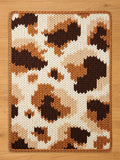 A rectangular, knitted chunky blanket with a brown border. The rug features a pattern of brown, tan, and cream-colored shapes, resembling a cowhide design. The blanket is made of a thick, textured yarn.