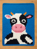 Chunky knit blanket featuring a cartoon cow design against a bright blue background. The blanket shows a black and white spotted cow with a pink snout and inner ears, small orange horns, and large stylized eyes, knit from thick, textured yarn.