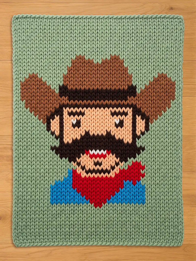 A knitted blanket featuring a pixelated cowboy design. The cowboy has a brown hat, beard, and mustache, a red bandana, and a blue shirt. The background is a light green color.