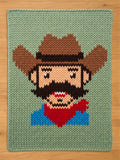 A knitted blanket featuring a pixelated cowboy design. The cowboy has a brown hat, beard, and mustache, a red bandana, and a blue shirt. The background is a light green color.