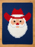 A knitted Christmas-themed square featuring a Santa Claus face. The design includes a red hat and beard, a peach-colored face, and a navy blue background. The Santa face is centered on the square.